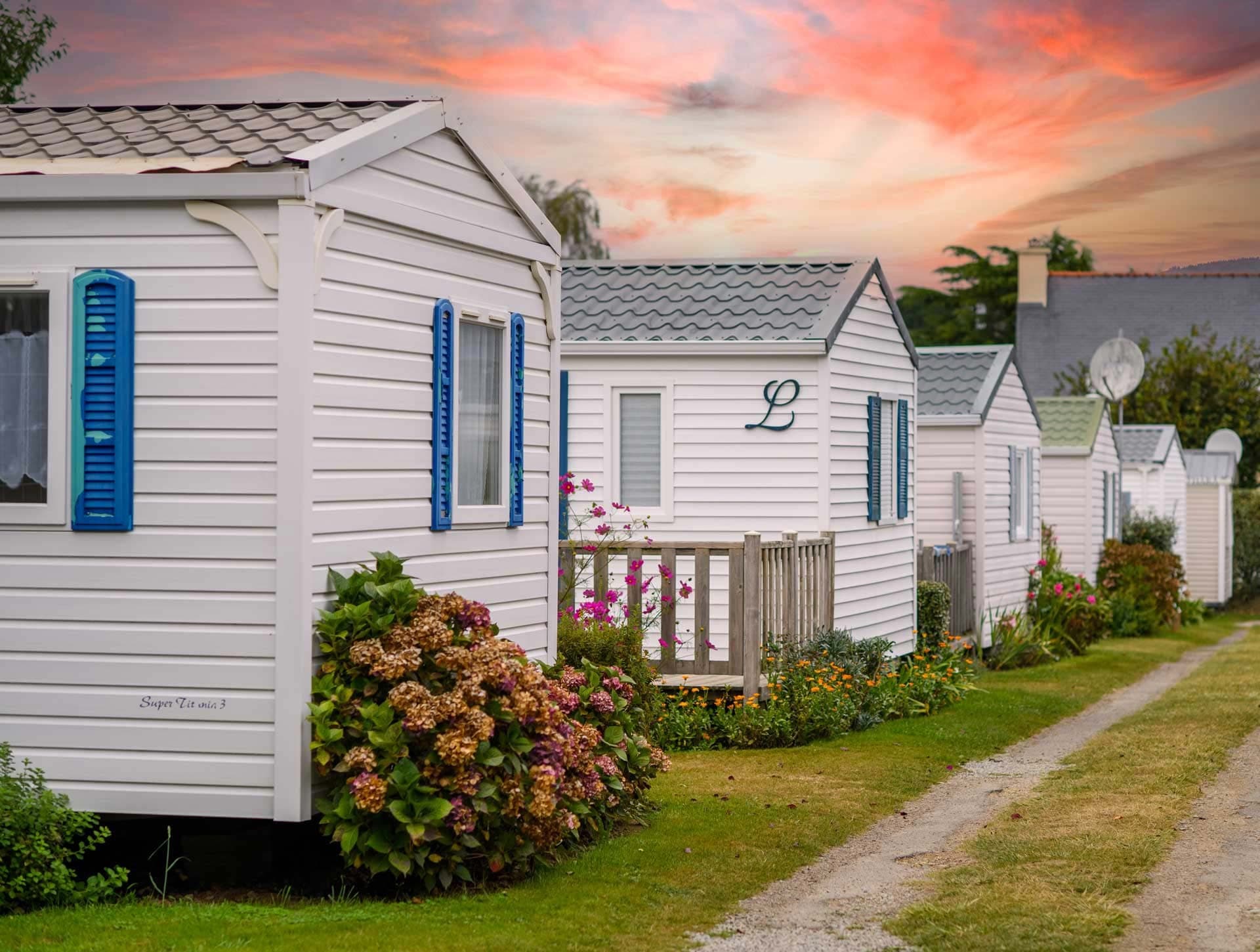 Alignement de mobil-homes blancs aux volets bleus dans un camping français, hortensias en pots et ciel pastel au coucher du soleil
